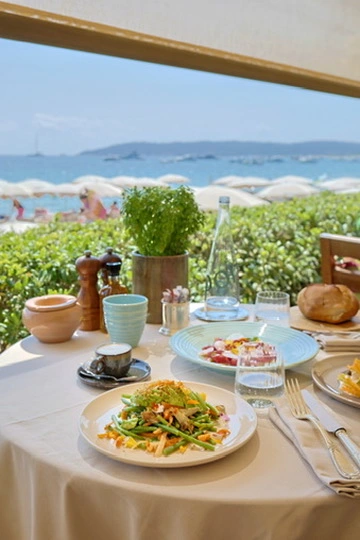 A beautiful outdoor table setting overlooking a sunny Mediterranean beach, featuring fresh meals beautifully presented on handcrafted tableware by Kaya Ceramics.
