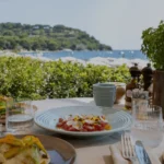 A sunlit outdoor table setting featuring fresh food plated on Kaya Ceramics' handmade Saint-Tropez collection, overlooking a beautiful Mediterranean beach.